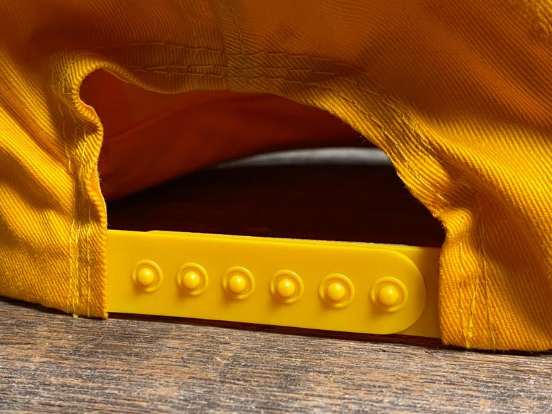 May include: Close-up of a yellow baseball cap, showing the adjustable strap and the underside of the brim. The cap is made of a textured fabric and has a bright yellow color. The adjustable strap is also yellow.