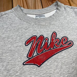 Puede incluir: Sudadera gris de cuello redondo con el logotipo de Nike en escritura roja con contorno blanco. El logotipo est&aacute; sobre un swoosh rojo. Peque&ntilde;o logotipo de Nike en el pecho.