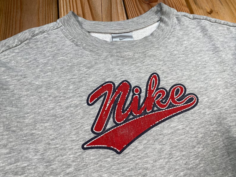 Puede incluir: Sudadera gris de cuello redondo con el logotipo de Nike en escritura roja con contorno blanco. El logotipo est&aacute; sobre un swoosh rojo. Peque&ntilde;o logotipo de Nike en el pecho.