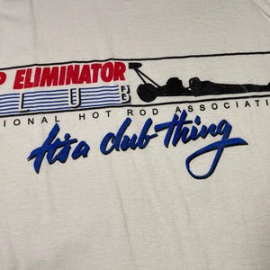 May include: White tank top with the text "TOP ELIMINATOR CLUB" in red and blue, with a black graphic of a dragster and the words "It's a club thing" in blue script. The words "NATIONAL HOT ROD ASSOCIATION" are also printed on the shirt.