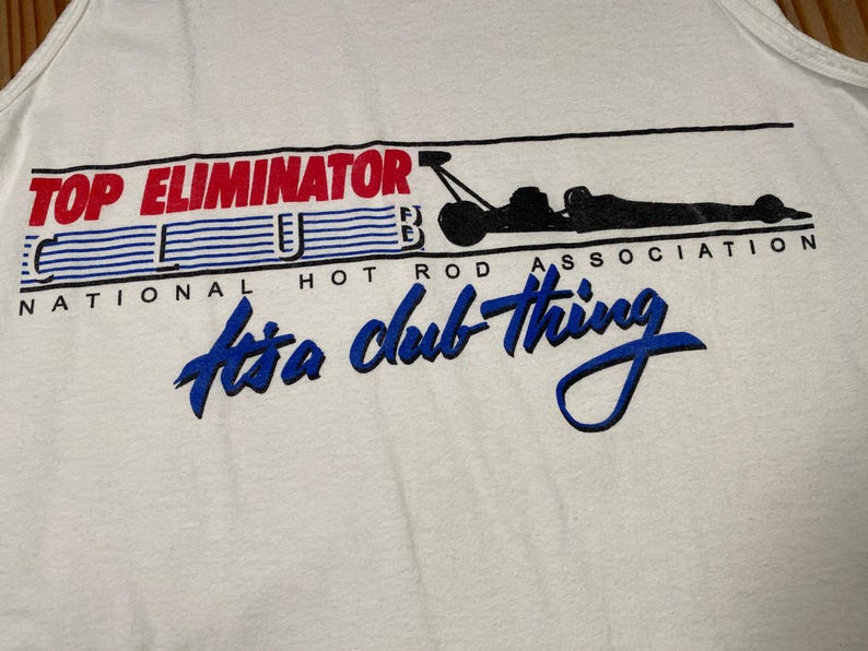May include: White tank top with the text "TOP ELIMINATOR CLUB" in red and blue, with a black graphic of a dragster and the words "It's a club thing" in blue script. The words "NATIONAL HOT ROD ASSOCIATION" are also printed on the shirt.