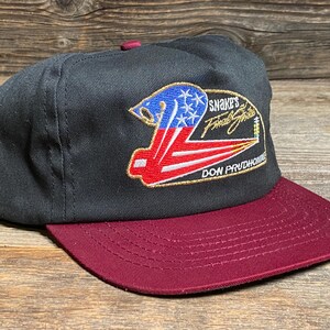 May include: Black and burgundy baseball cap with a flat peak. The cap has an embroidered design with the text "Snake's Final Strike" and "Don Prudhomme". The design incorporates red, white, and blue elements.