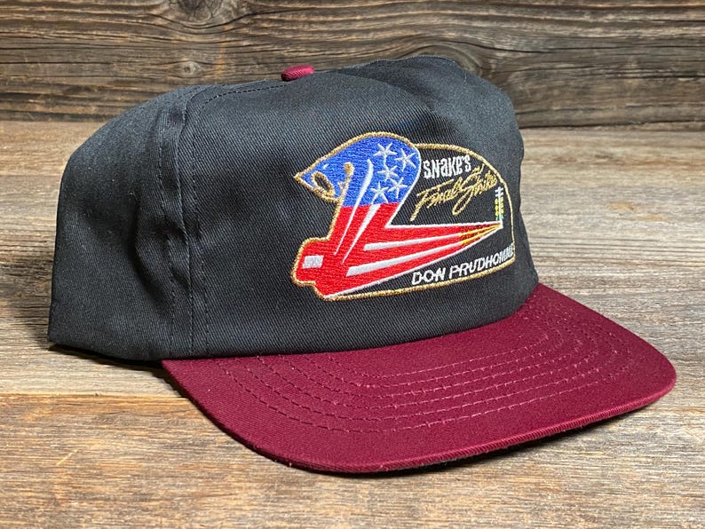 May include: Black and burgundy baseball cap with a flat peak. The cap has an embroidered design with the text "Snake's Final Strike" and "Don Prudhomme". The design incorporates red, white, and blue elements.