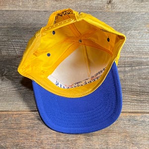 May include: A yellow and blue baseball cap with a flat brim. The cap has a yellow crown and a blue brim. The interior is white with printed text. The cap is made of a durable material and has an adjustable snap closure.