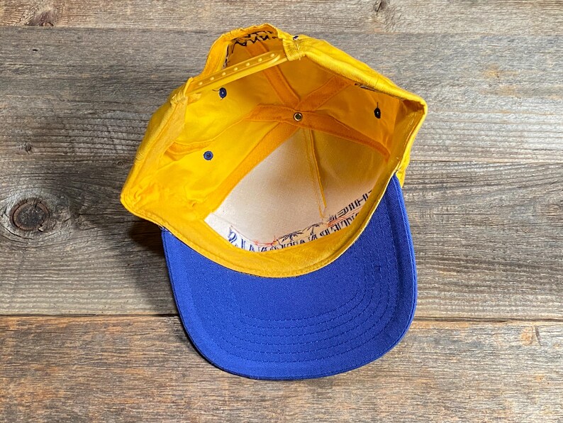 May include: A yellow and blue baseball cap with a flat brim. The cap has a yellow crown and a blue brim. The interior is white with printed text. The cap is made of a durable material and has an adjustable snap closure.