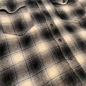 May include: Close-up of a black, white, and grey plaid flannel shirt. The shirt has two chest pockets and black buttons down the front and on the cuffs. The fabric has a soft, textured appearance.