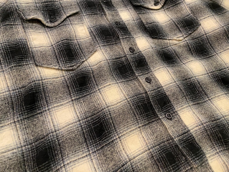 May include: Close-up of a black, white, and grey plaid flannel shirt. The shirt has two chest pockets and black buttons down the front and on the cuffs. The fabric has a soft, textured appearance.