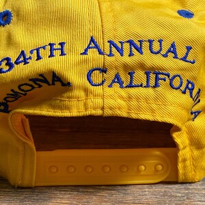 May include: Yellow baseball cap with blue embroidered text that reads "34th Annual Pomona California". The cap has a yellow adjustable snap closure and blue eyelets.