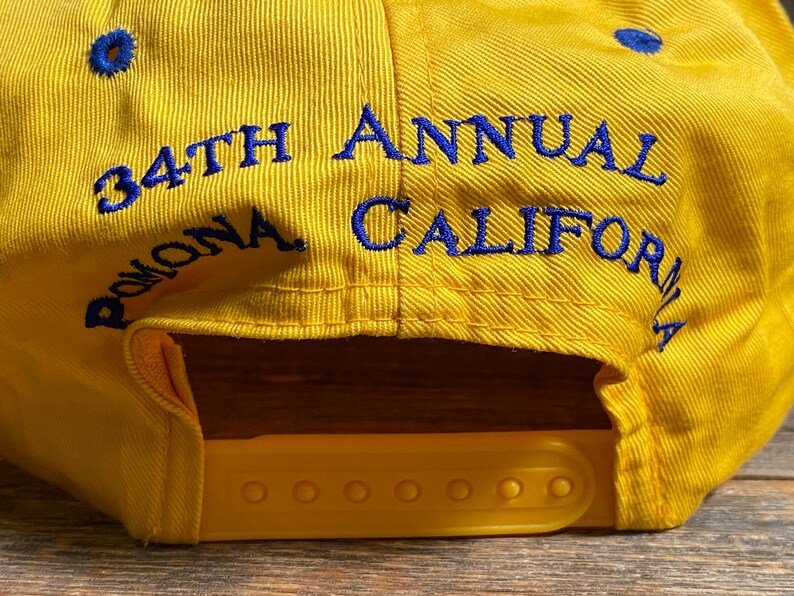 May include: Yellow baseball cap with blue embroidered text that reads "34th Annual Pomona California". The cap has a yellow adjustable snap closure and blue eyelets.