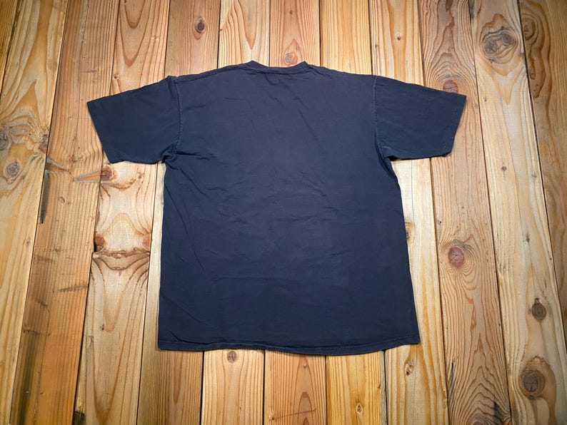 Puede incluir: Una camiseta de manga corta de color azul marino oscuro s&oacute;lido con cuello redondo. La prenda se muestra extendida, revelando la parte posterior lisa de la camiseta sin gr&aacute;ficos, patrones ni texto visibles en la superficie de la tela.