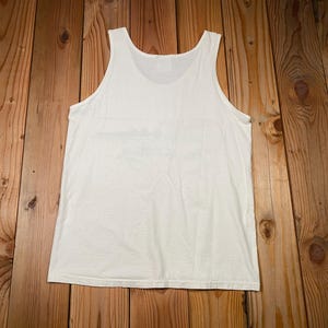 May include: A white tank top made of a soft material. The sleeveless shirt is laid flat on a wooden surface. The top has a scoop neck and a simple design.