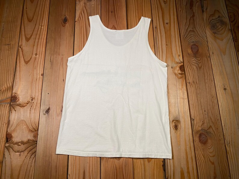 May include: A white tank top made of a soft material. The sleeveless shirt is laid flat on a wooden surface. The top has a scoop neck and a simple design.