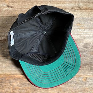 May include: Black baseball cap with a green peak and red stitching. The underside of the cap is black with a mesh panel. The cap is on a wooden surface.