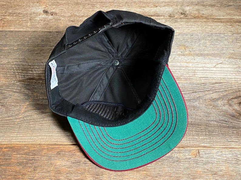 May include: Black baseball cap with a green peak and red stitching. The underside of the cap is black with a mesh panel. The cap is on a wooden surface.