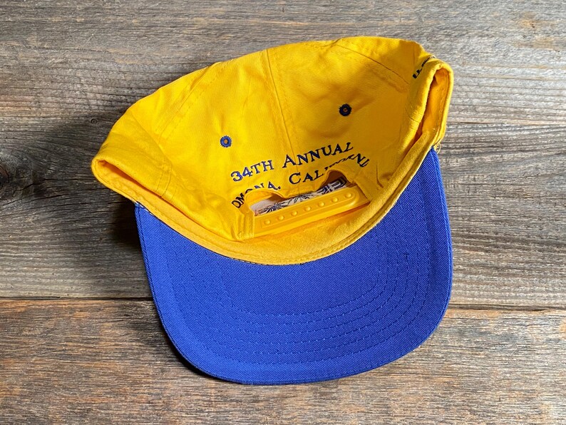 May include: A yellow and blue baseball cap with the text "34th Annual Omona, CA" visible. The cap has a yellow brim and a blue bill. The cap is made of a durable fabric and has an adjustable snap closure.