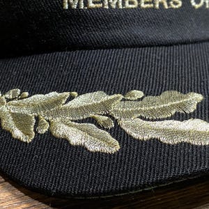 May include: Black baseball cap with gold embroidered leaf design on the side. The text "MEMBERS ONLY" is embroidered in gold on the front of the cap. The cap is made of a textured fabric.