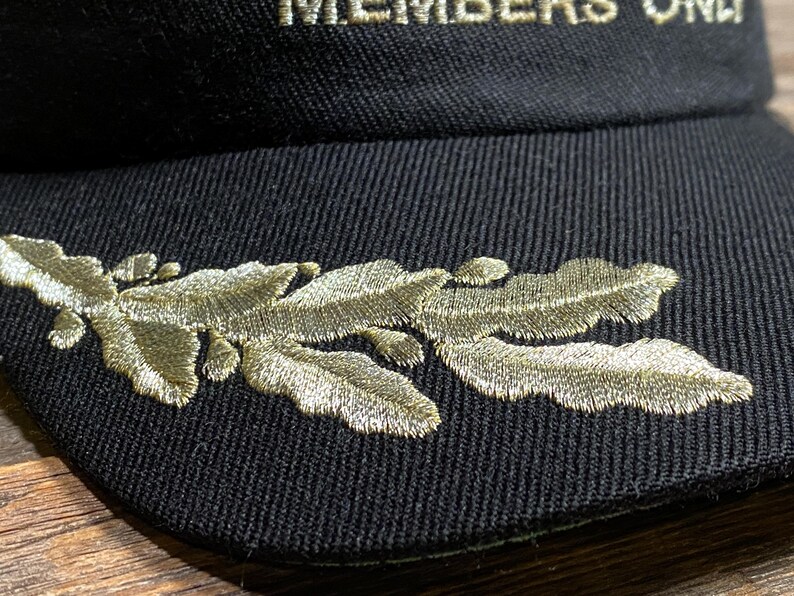 May include: Black baseball cap with gold embroidered leaf design on the side. The text "MEMBERS ONLY" is embroidered in gold on the front of the cap. The cap is made of a textured fabric.