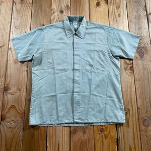 May include: A light sage green short-sleeve button-down shirt featuring a classic collar, a front button placket, and two symmetrical chest pockets. The shirt is constructed with a straight hemline and visible stitching along the sleeves and pockets.