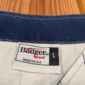 Peut inclure: Un maillot de baseball blanc &agrave; fines rayures bleu marine avec un col bleu marine. Une &eacute;tiquette en tissu blanc est cousue &agrave; l'encolure avec le texte Badger Sport, la lettre L et la mention MADE IN U.S.A. en lettres noires et rouges.