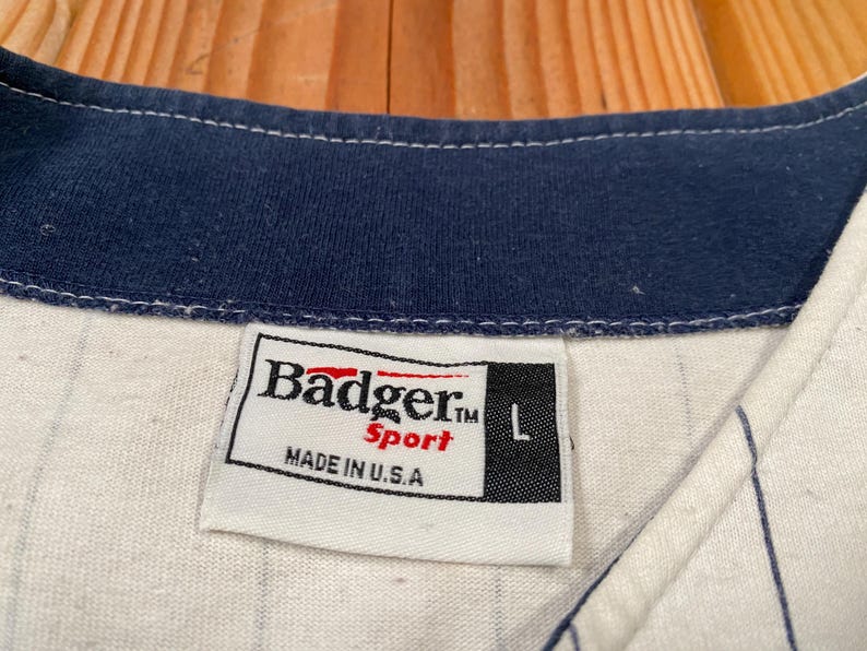 Peut inclure: Un maillot de baseball blanc &agrave; fines rayures bleu marine avec un col bleu marine. Une &eacute;tiquette en tissu blanc est cousue &agrave; l'encolure avec le texte Badger Sport, la lettre L et la mention MADE IN U.S.A. en lettres noires et rouges.