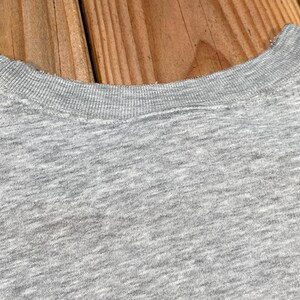 Puede incluir: Primer plano de una camiseta gris con cuello redondo. El cuello de la camisa es acanalado y la tela tiene una textura jaspeada. El fondo es una superficie de madera.