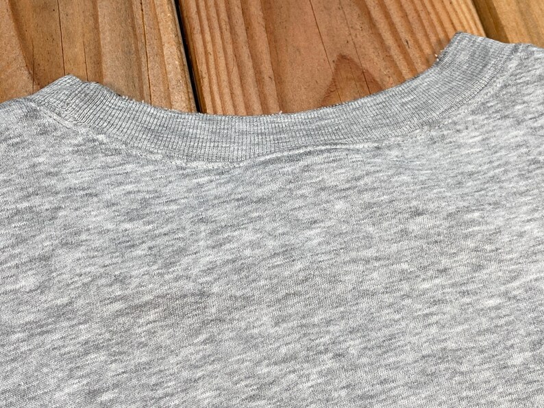 Puede incluir: Primer plano de una camiseta gris con cuello redondo. El cuello de la camisa es acanalado y la tela tiene una textura jaspeada. El fondo es una superficie de madera.