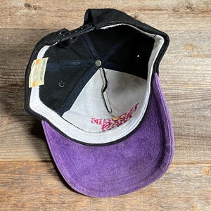 May include: Black baseball cap with a purple suede bill. The cap has a black adjustable strap and a white interior with pink text. A small tag is attached to the side.