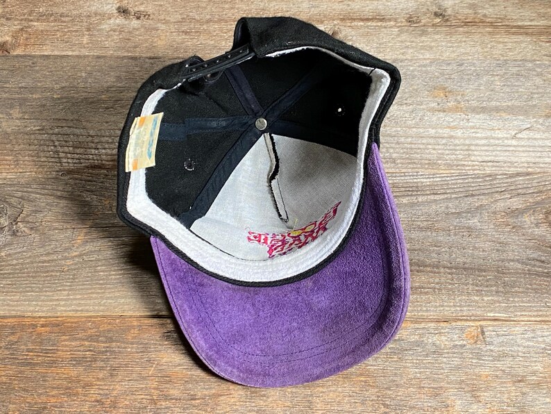 May include: Black baseball cap with a purple suede bill. The cap has a black adjustable strap and a white interior with pink text. A small tag is attached to the side.