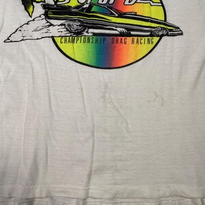 Pu&ograve; includere: T-shirt bianco con grafica colorata di un'auto da drag racing con la scritta "CHAMPIONSHIP DRAG RACING". L'auto &egrave; nera e gialla, su uno sfondo arcobaleno.