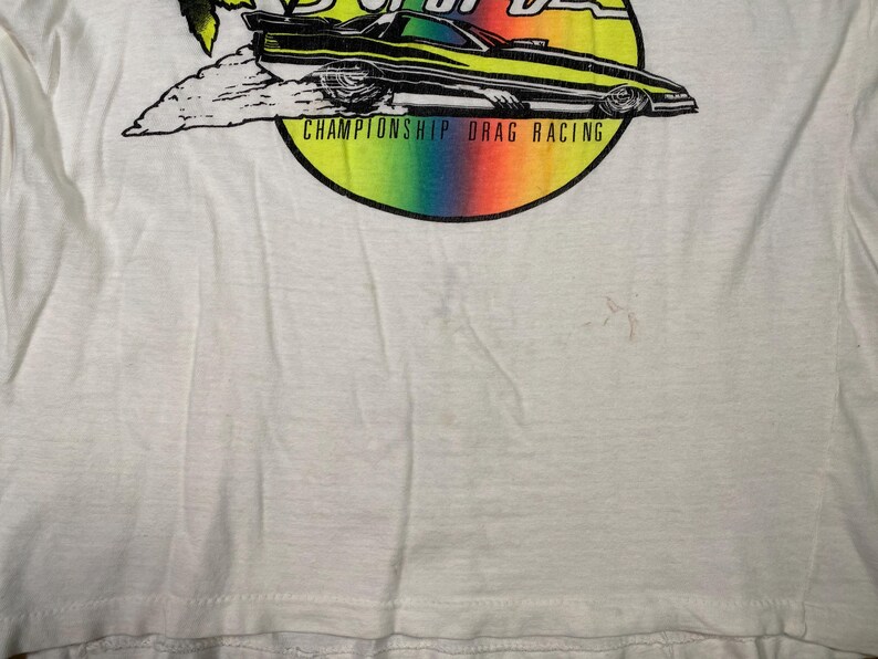 Pu&ograve; includere: T-shirt bianco con grafica colorata di un'auto da drag racing con la scritta "CHAMPIONSHIP DRAG RACING". L'auto &egrave; nera e gialla, su uno sfondo arcobaleno.