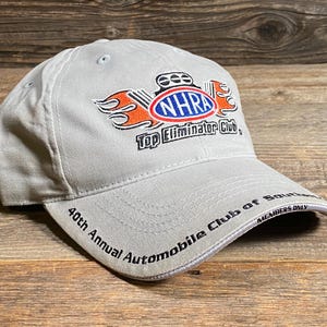 May include: A light grey baseball cap with the NHRA Top Eliminator Club logo in red, orange, and blue. The cap also displays "40th Annual Automobile Club of Southern California" and "Members Only" text.