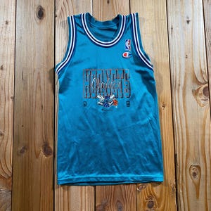 May include: A teal Charlotte Hornets basketball jersey featuring a white and purple striped trim on the collar and armholes. The front displays the word Hornets in block letters, the script word Charlotte, and a hornet logo with a basketball.