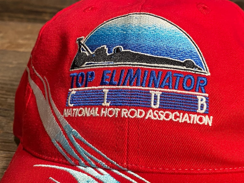 Op de afbeelding: Een felrode baseballpet met een geborduurd logo met een blauwe gradi&euml;nt halve cirkel, een zwart silhouet van een raceauto en de tekst TOP ELIMINATOR CLUB NATIONAL HOT ROD ASSOCIATION. Een lichtblauw en wit swoosh-ontwerp siert de zijkant.