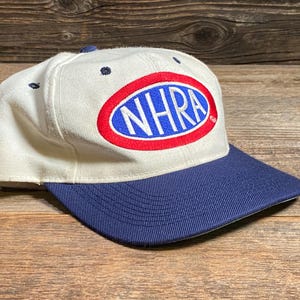 Op de afbeelding: Een witte baseballcap met een marineblauwe klep. De cap heeft het NHRA-logo in een rode en blauwe ovaal op de voorkant. De hoed is gemaakt van een duurzame stof en heeft een klassiek, gestructureerd ontwerp.