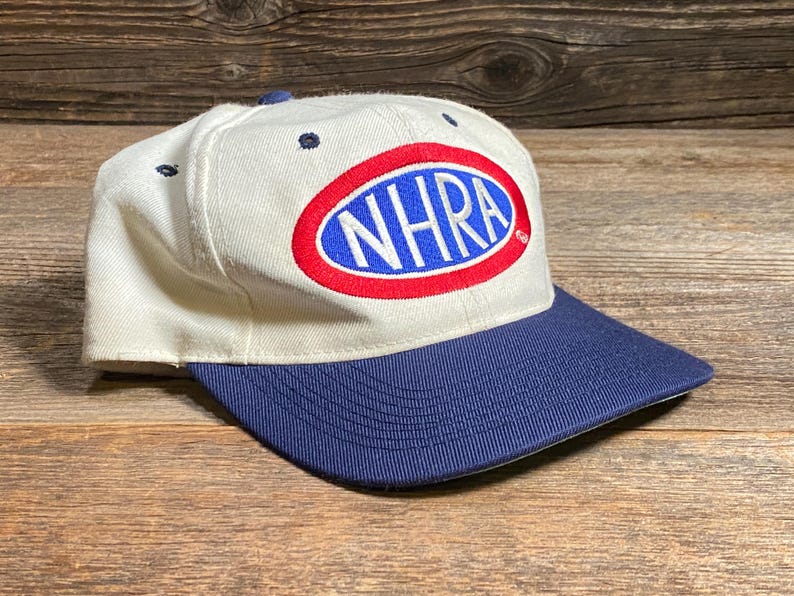 Op de afbeelding: Een witte baseballcap met een marineblauwe klep. De cap heeft het NHRA-logo in een rode en blauwe ovaal op de voorkant. De hoed is gemaakt van een duurzame stof en heeft een klassiek, gestructureerd ontwerp.
