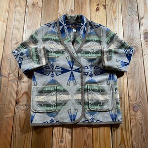 May include: A long-sleeve jacket featuring a shawl collar and two front patch pockets. The garment is crafted from a grey fabric adorned with a geometric pattern in shades of blue, green, and cream, finished with a brown trim along the edges.