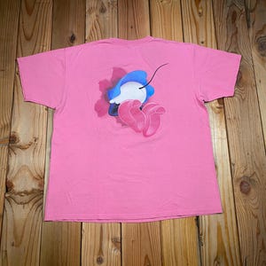 May include: A short-sleeved pink t-shirt featuring a central graphic on the back. The design depicts a stylized blue and white rounded shape with a thin black line extending upward, positioned above two overlapping pink petal-like forms.