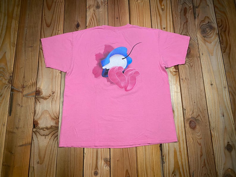 May include: A short-sleeved pink t-shirt featuring a central graphic on the back. The design depicts a stylized blue and white rounded shape with a thin black line extending upward, positioned above two overlapping pink petal-like forms.