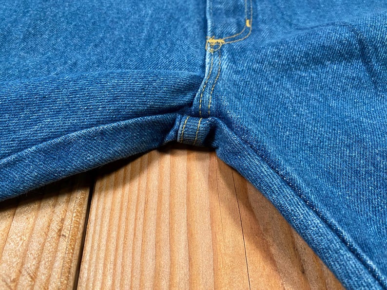 May include: Close-up of blue denim jeans. The jeans have yellow stitching and are on a wooden surface. The fabric is a medium wash denim, showing the texture of the material.
