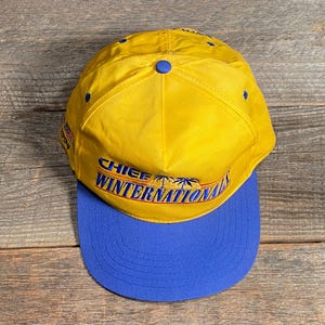 May include: Yellow and blue baseball cap with the text "CHIEF WINTERNATIONAL" across the front. The cap has a curved brim and a button on top. The hat is made of a durable fabric.
