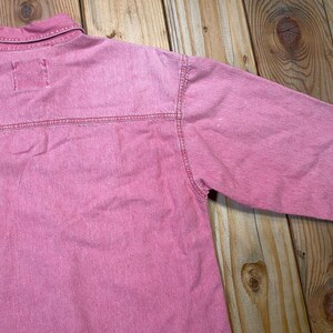 Puede incluir: Una camisa de manga larga en un tono rosa deste&ntilde;ido, con cuello cl&aacute;sico, costura horizontal en el canes&uacute; trasero y detalles de costuras visibles a lo largo de los hombros y las mangas. La prenda est&aacute; confeccionada en un tejido texturizado.