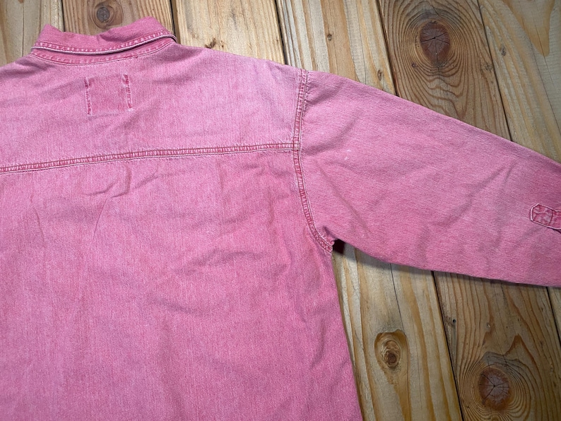 Puede incluir: Una camisa de manga larga en un tono rosa deste&ntilde;ido, con cuello cl&aacute;sico, costura horizontal en el canes&uacute; trasero y detalles de costuras visibles a lo largo de los hombros y las mangas. La prenda est&aacute; confeccionada en un tejido texturizado.