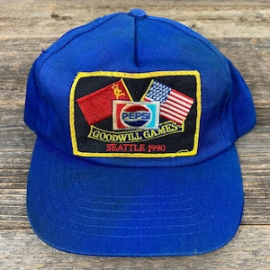 May include: A blue baseball cap featuring a patch with the Pepsi logo, flags, and text that reads "Goodwill Games Seattle 1990." The cap is made of a durable fabric and has a curved brim.