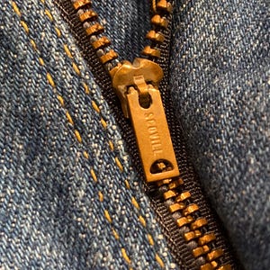 May include: Close-up of a blue denim fabric with a brass zipper. The zipper pull is marked with the word "SCOVILL". The denim has yellow stitching.