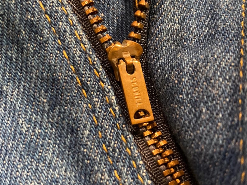 May include: Close-up of a blue denim fabric with a brass zipper. The zipper pull is marked with the word "SCOVILL". The denim has yellow stitching.