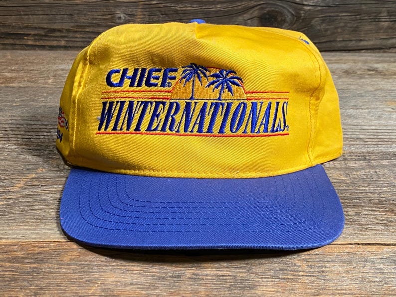 May include: A yellow and blue baseball cap with the text "CHIEF WINTERNATIONALS" embroidered on the front. The cap features a blue brim and a yellow crown. The text is in blue and red, with palm tree graphics.