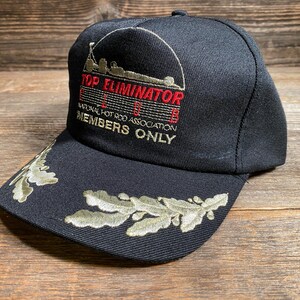 May include: Black baseball cap with embroidered text "TOP ELIMINATOR" and "NATIONAL HOT ROD ASSOCIATION MEMBERS ONLY". The brim features olive green leaf embroidery. The cap is made of a durable fabric.