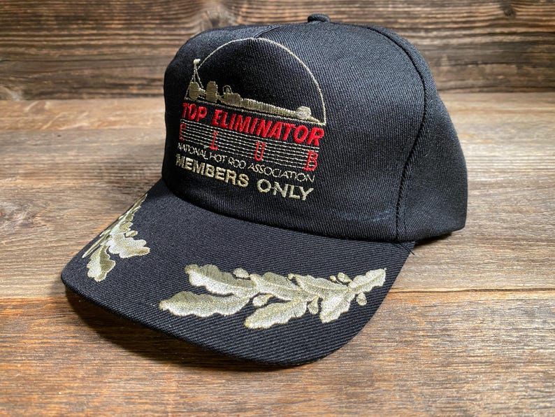 May include: Black baseball cap with embroidered text "TOP ELIMINATOR" and "NATIONAL HOT ROD ASSOCIATION MEMBERS ONLY". The brim features olive green leaf embroidery. The cap is made of a durable fabric.