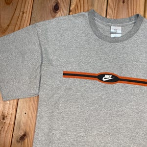 Puede incluir: Camiseta gris con cuello redondo y mangas cortas. La camiseta presenta un dise&ntilde;o de rayas horizontales naranja y negro en el pecho, con el logotipo blanco de Nike en el centro. El logotipo de Nike tambi&eacute;n es visible en la etiqueta.