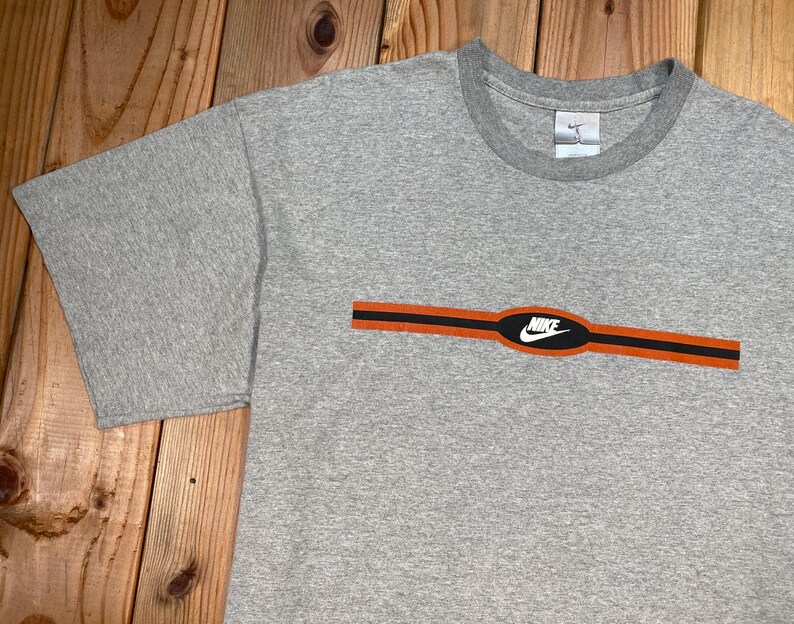 Puede incluir: Camiseta gris con cuello redondo y mangas cortas. La camiseta presenta un dise&ntilde;o de rayas horizontales naranja y negro en el pecho, con el logotipo blanco de Nike en el centro. El logotipo de Nike tambi&eacute;n es visible en la etiqueta.